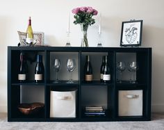 a shelf with wine glasses, bottles and other items on it in front of a white wall