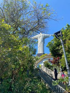 the statue of christ is on top of a hill near trees and stairs with people walking up it