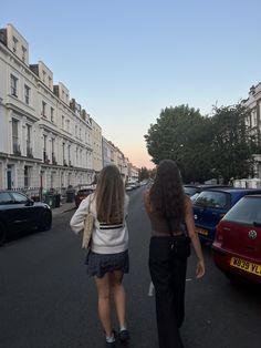 two women walking down the street in front of parked cars and tall white buildings on both sides