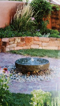 a water fountain in the middle of a garden with rocks and grass around it, surrounded by flowers