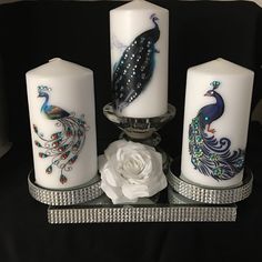 three white candles with peacocks on them sitting next to a flower and candle holder