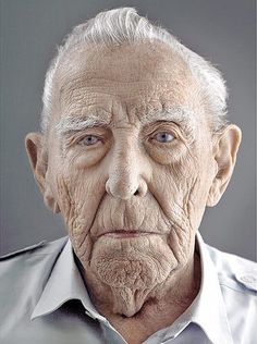 an old man with white hair and blue eyes looks at the camera while wearing a gray shirt