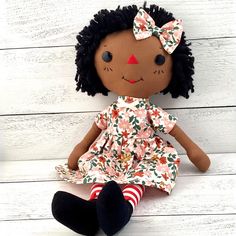 a doll with black hair wearing a floral dress and striped socks sits on a white wooden background