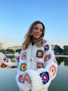 a woman standing in front of water wearing a white crocheted jacket with multicolored circles on it