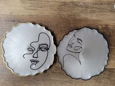 two plates with drawings on them sitting on a wooden table next to each other, one has a woman's face painted on it