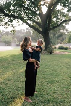 a woman holding a baby in her arms while standing on the grass near a tree