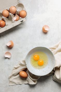 two eggs in a white bowl next to an egg carton on a gray surface