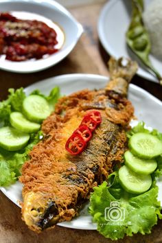 a white plate topped with fish next to cucumbers and sauce on top of rice