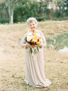 an older woman holding a bouquet of flowers in front of a body of water with trees in the background