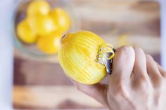 a hand holding a peeled lemon in front of a glass bowl with yellow fruit behind it