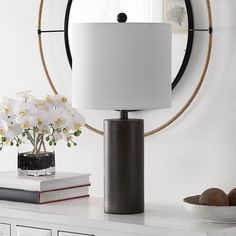 a table with a lamp and flowers on it in front of a round wall mirror