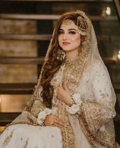 a woman in a white and gold bridal gown sitting on a bench with her hands clasped