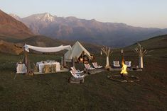 a tent set up in the middle of a field with mountains in the background and chairs around it