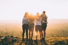 four people standing on top of a hill with their arms around each other as the sun goes down