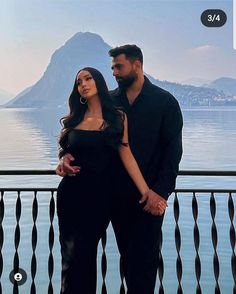 a man and woman standing next to each other in front of a lake with mountains