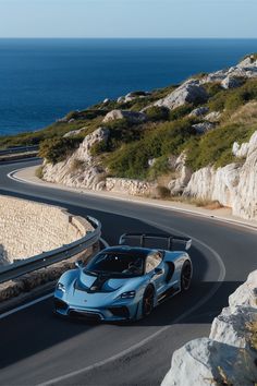 a blue sports car driving down a road next to the ocean in front of some rocks