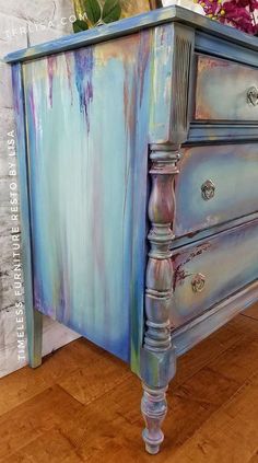 an old dresser painted in blue and purple with flowers sitting on it's top