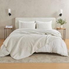 a bed with white sheets and pillows in a room next to a plant on a table