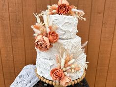 a three tiered cake decorated with flowers and feathers on a lace doily next to a wooden wall