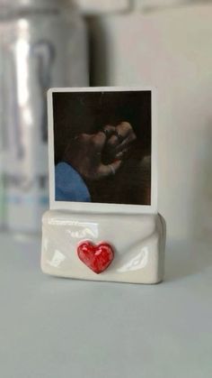 a small white photo frame with a red heart on it and a hand holding a finger