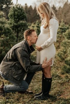 a pregnant man kneeling down to kiss his pregnant wife's belly in the woods