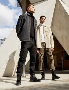 two young men standing next to each other in front of a white building, one wearing black boots