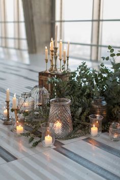 a table topped with lots of candles and vases filled with greenery on top of a wooden floor