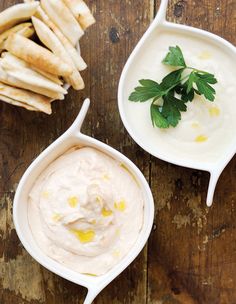 two bowls filled with dip next to some pita bread on a wooden table,