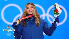 a woman holding flowers and a medal in front of a blue wall with the olympic rings behind her