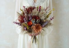 a woman wearing a white dress holding a bouquet of dried flowers and foliage in her hands