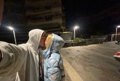 a person in a hooded jacket kissing a child on the street at night with lights behind them