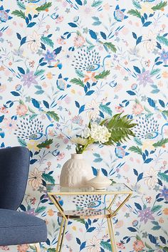 a blue chair sitting next to a white vase with flowers on it in front of a floral wallpaper