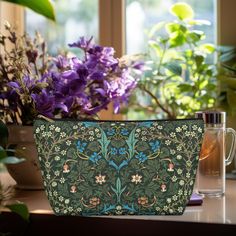 a green and blue purse sitting on top of a table next to purple flowers in front of a window