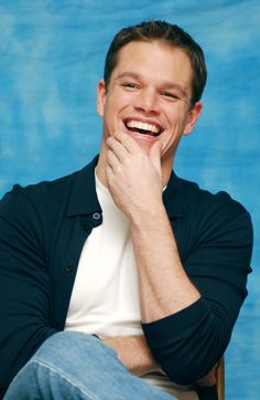 a smiling man sitting in a chair with his hand on his chin