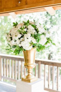 a gold vase filled with white flowers sitting on top of a wooden table next to a railing