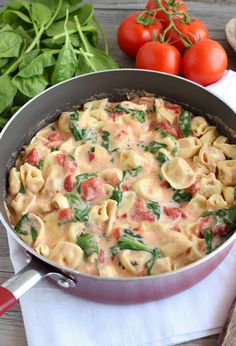 a pan filled with pasta and spinach on top of a wooden table next to tomatoes