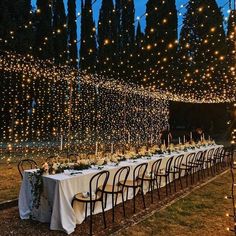 a long table is set up with white linens and lite - up trees in the background