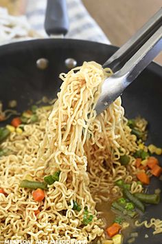 noodles being cooked in a wok with tongs on the side and vegetables mixed into it