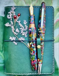 two pens sitting on top of a green leather case next to leaves and berries in it