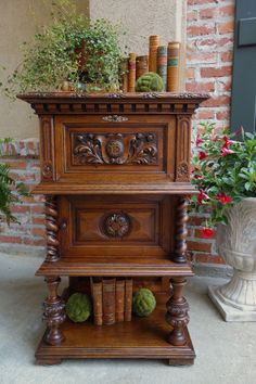 an old wooden cabinet with books and plants on top in front of a brick wall