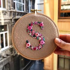 a hand holding up a embroidery hoop with the letter s made out of flowers