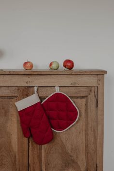 two red oven mitts hanging from a wooden cabinet next to an apple on top of it