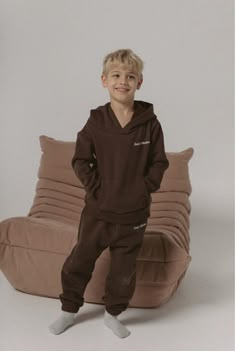 a young boy standing in front of a couch wearing a brown sweatshirt and sweatpants