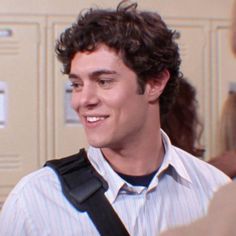 a young man with curly hair and suspenders smiles at the camera while standing in front of lockers