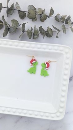 the small green dinosaur earrings have santa's hats on them and are sitting on a white tray