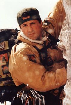 a man with a backpack on his back standing in front of a rock wall and looking at the camera