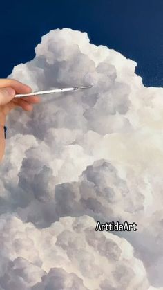 a hand holding a paintbrush in front of some clouds