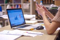 a woman sitting at a desk using her cell phone and laptop computer with papers on the table next to her