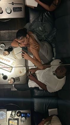 two people sitting at a table looking at their cell phones while they are surrounded by other people