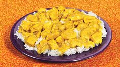 a purple plate topped with rice covered in yellow chicken and white sauce on an orange tablecloth